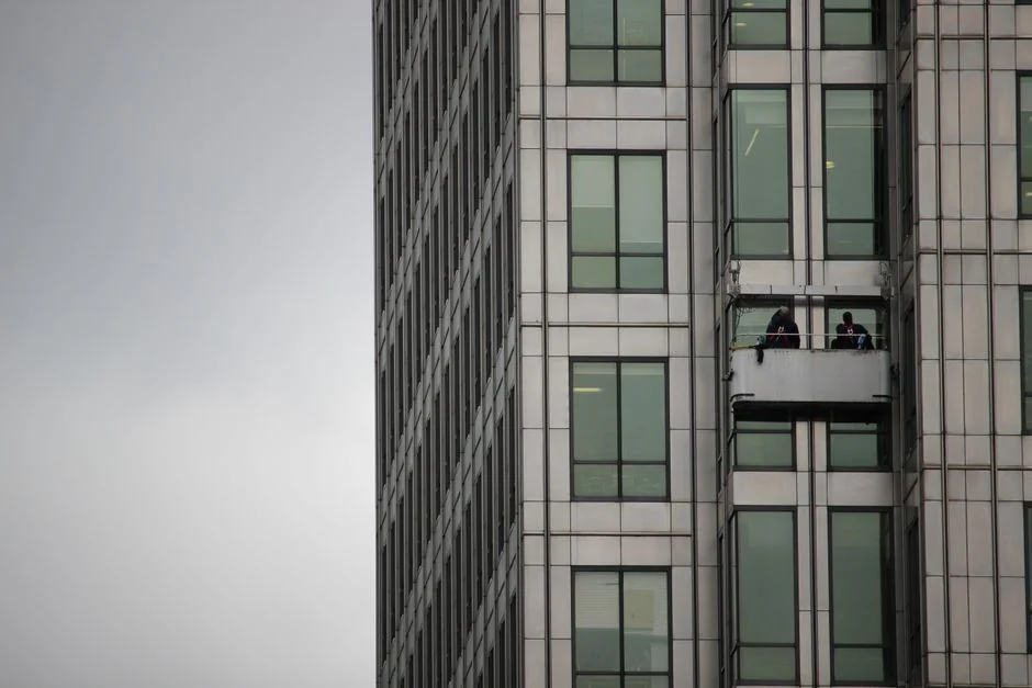 our-story Two workers clean windows on a modern glass building facade using a suspended platform.